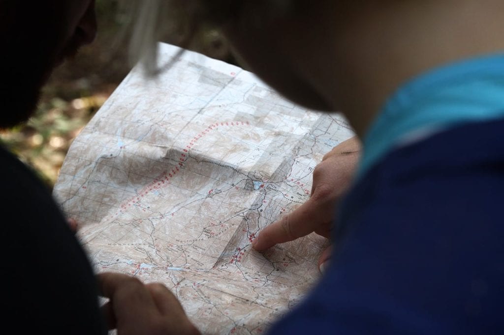 Photo of two people looking at a topographical map. The person on the right is pointing at something in the middle of the map. The background looks like they're in the woods.